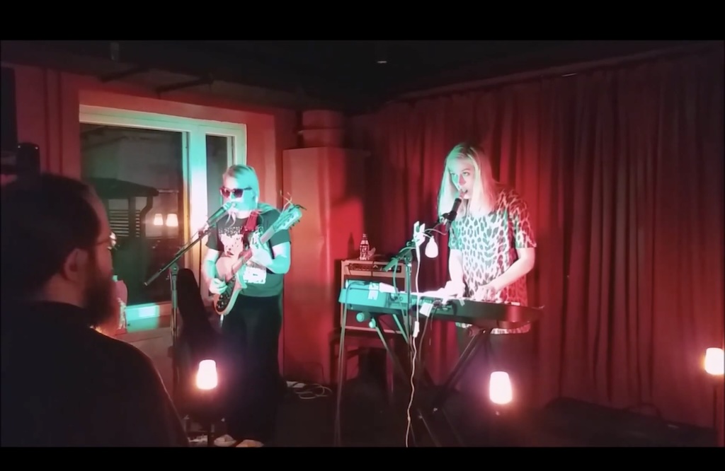 Image of band performing in front of red curtain, next to window and red wall, playing guitar and keyboard and wearing leopard print and band tshirts