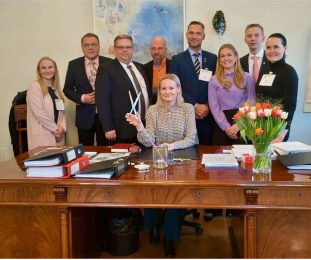 Picture of blonde woman in business blouse at a desk holding a large pair of scissors and smiling while people in business attire stand behind her